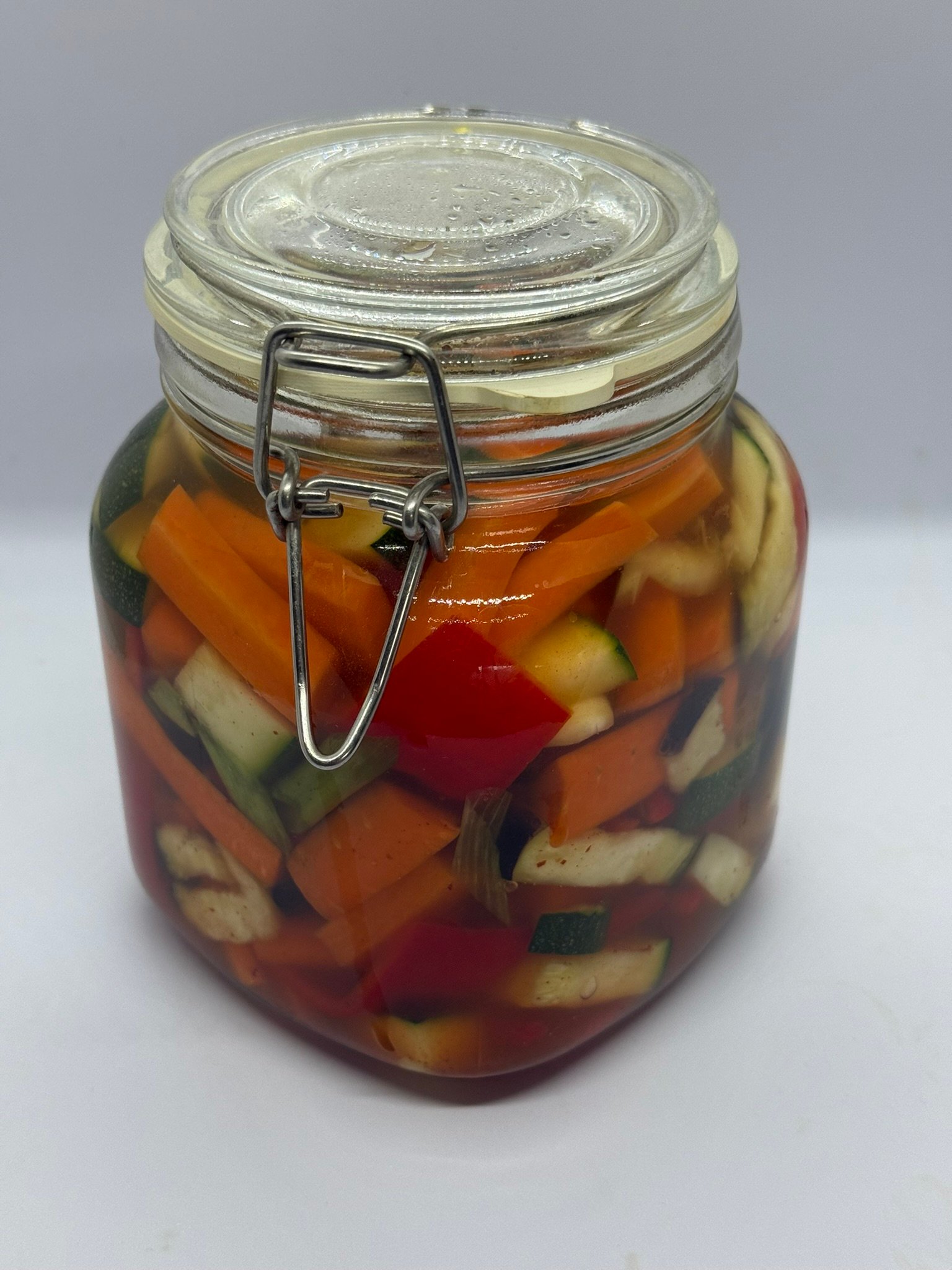 Een glazen pot gevuld met kleurrijke stukjes courgette, paprika en knoflook in een troebele zoutoplossing.