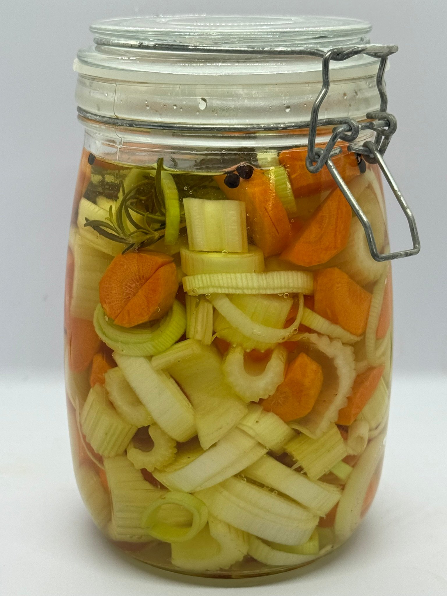 Verschillende lagen kleurrijke groenten zoals radijs, bloemkool en wortel in een glazen pot met heldere inmaakvloeistof.